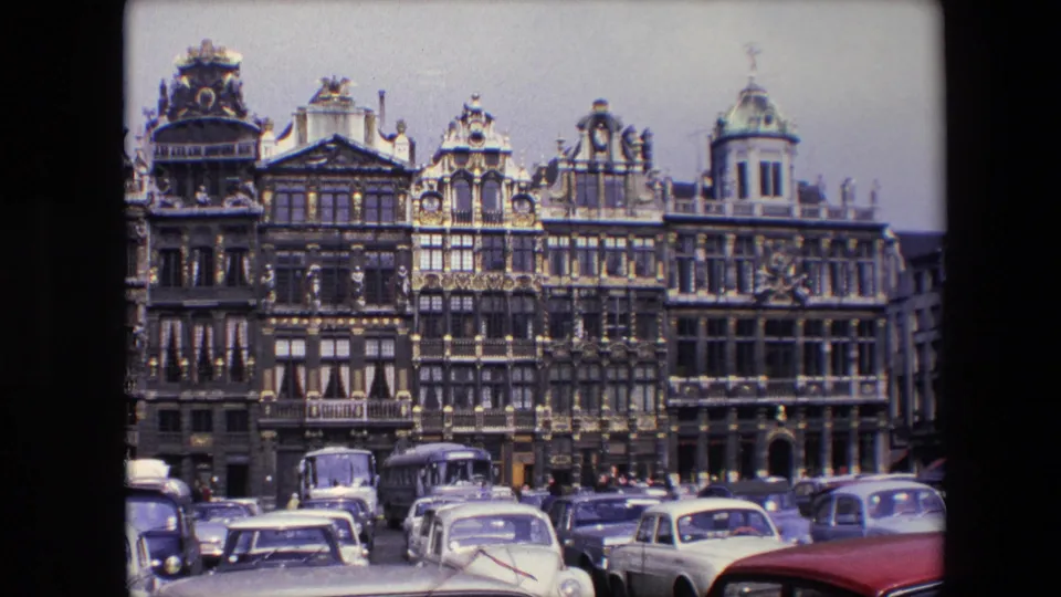 1967 Brussels home-movie frame showing ornate historic facades with gold-toned detailing above a foreground of parked cars and street traffic.