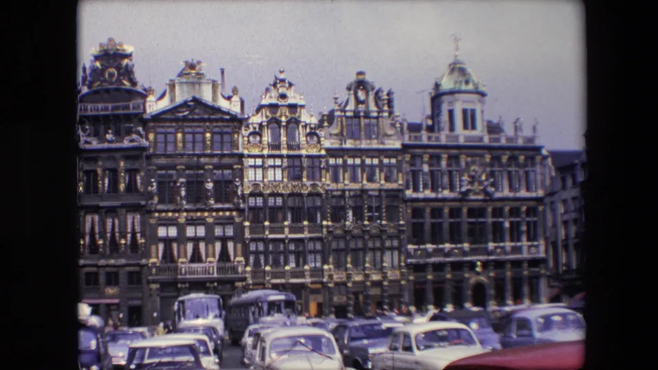 1967 Brussels frame showing a visitor's view across parked cars toward ornate multistory buildings with decorative gables and gilded details.