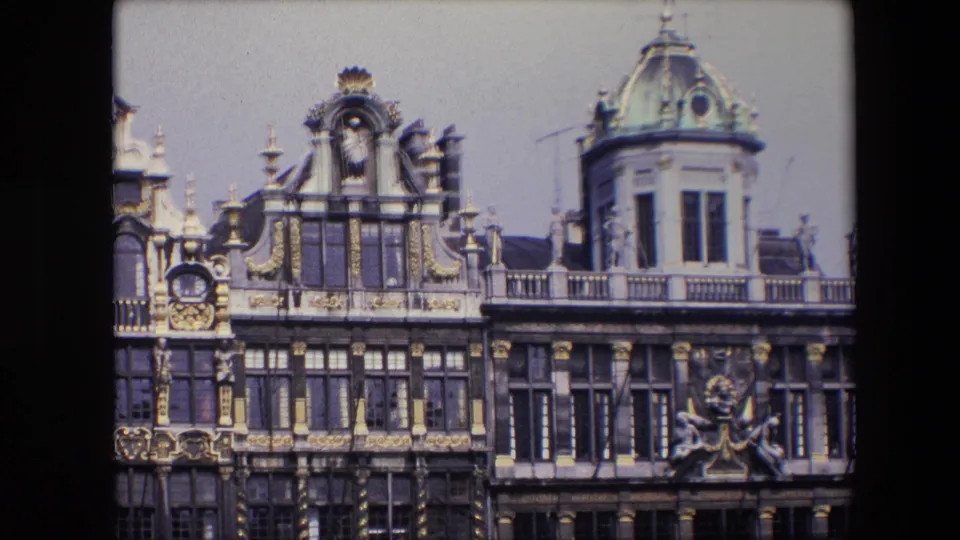 1967 8mm Brussels frame focusing on upper stories of historic facades, with gilded trim, dark masonry, tall windows, and a domed turret at right.
