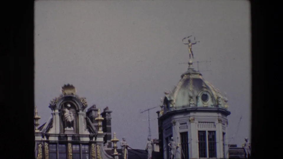 1967 Brussels home-movie frame with the camera framing the upper facade, gilded roof ornament, classical sculpture niches, rooftop antennas, and the domed tower at right.