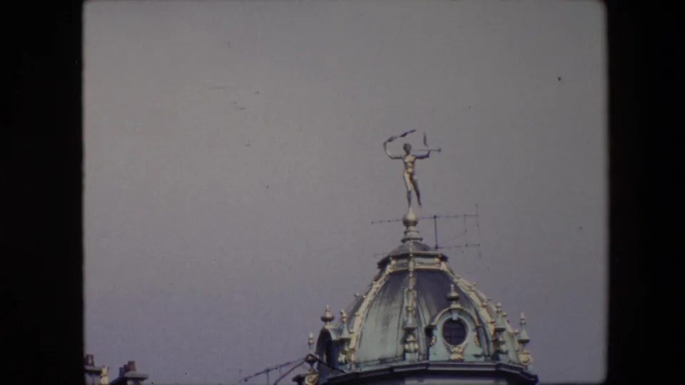 1967 Brussels frame showing the ornate dome cropped tighter, with a small golden statue above the roof, visible antennas, round dormer detail, and open pale sky.