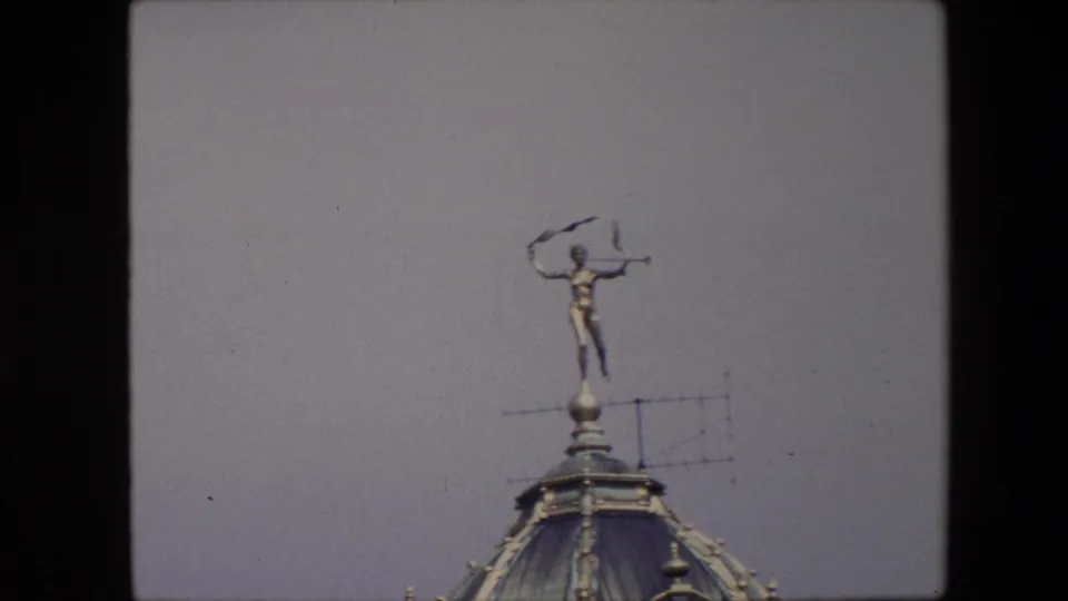 Handheld 8mm view from Brussels in 1967 showing the ornate dome, rooftop figure, and antenna framed below a pale sky.
