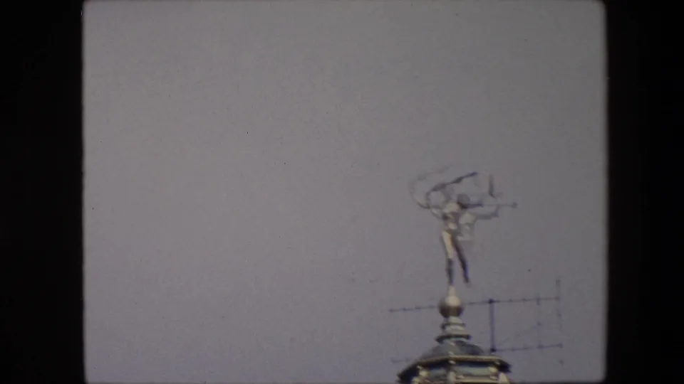 Softer 1967 Brussels 8mm frame with motion blur around the rooftop statue, dome edge, and nearby antenna against the sky.