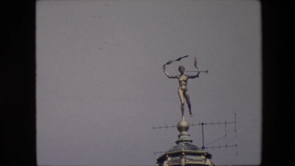 1967 Brussels footage frame showing a gold-toned nude male rooftop statue with a trumpet and flowing ribbon above a domed building detail and antenna.