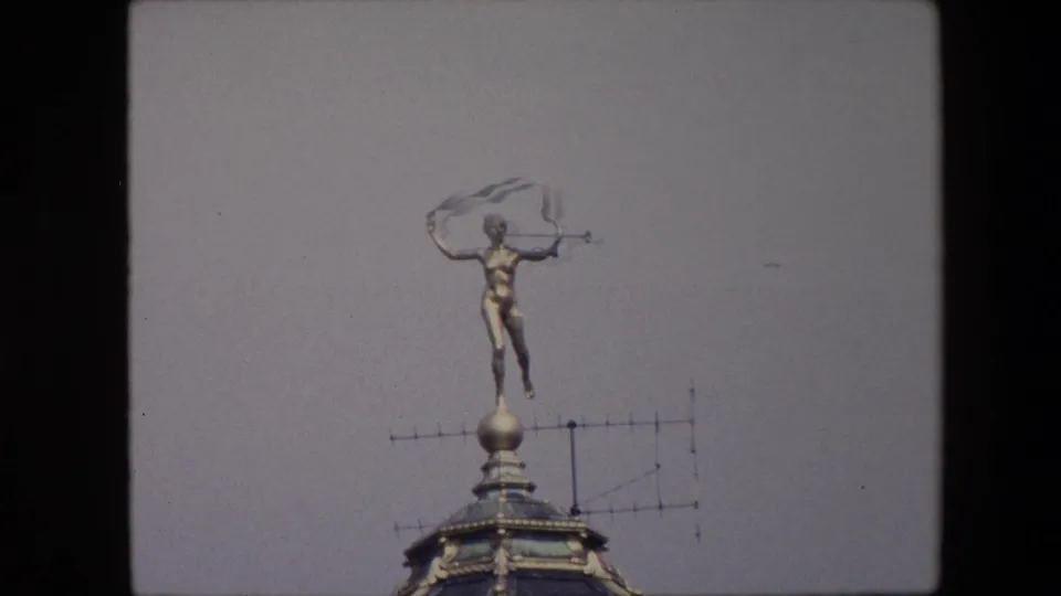 1967 Brussels frame with the gold-toned rooftop figure centered above a dome, holding a trumpet and ribbon near a television antenna.