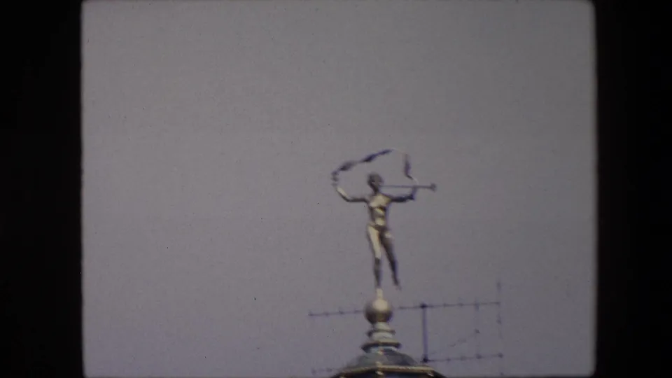 Slightly soft 1967 Brussels frame showing the rooftop statue with trumpet and ribbon above a domed building detail, with antenna lines crossing the background.