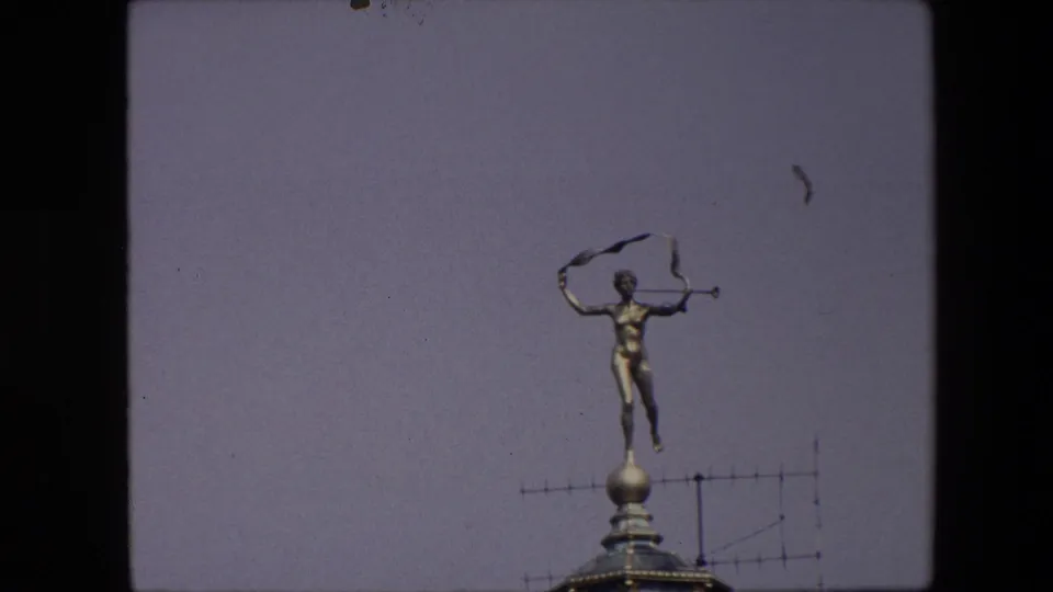 1967 Brussels frame with the gold-toned trumpet figure and ribbon on a rooftop finial, set against open sky with visible 8mm border darkness.