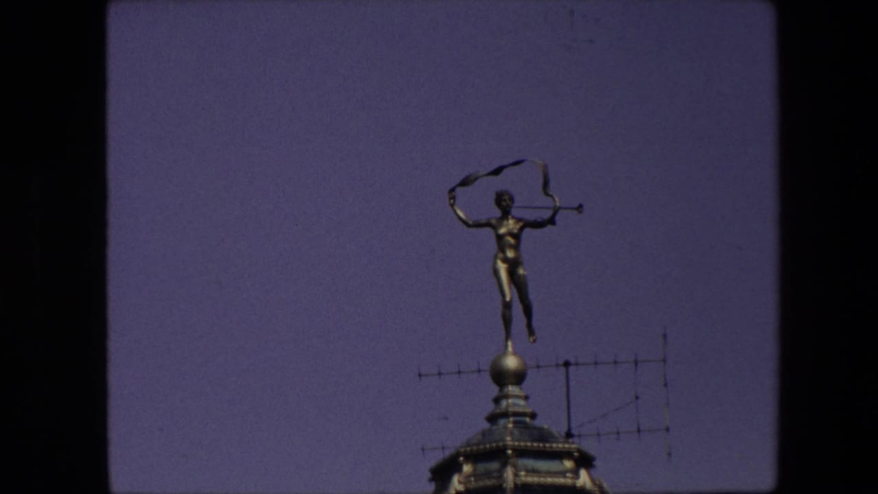 1967 Brussels 8mm frame showing a small golden rooftop figure holding a violin-like object above a weathervane or finial against a purple evening sky.