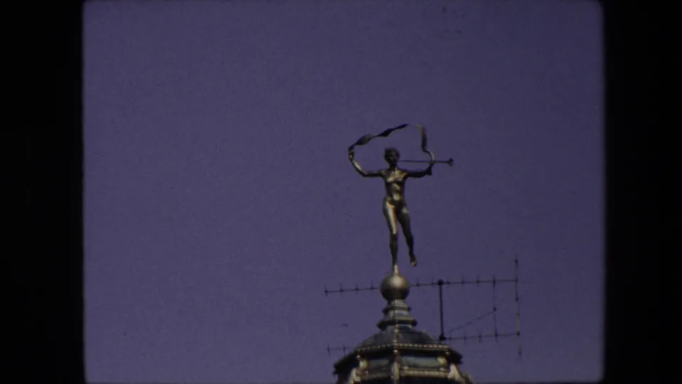 1967 Brussels 8mm frame showing a small golden rooftop figure holding a violin-like object above a weathervane or finial against a purple evening sky.