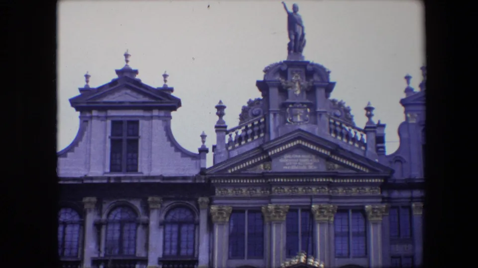 1967 Brussels home-movie frame showing a tighter view of decorated historic architecture, with roof ornament, arched windows, carved stonework, and a statue above the central roofline.