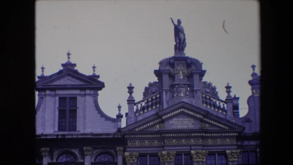 1967 Brussels frame centered on a rooftop statue and richly ornamented roofline, with adjacent gables, stone finials, arched windows, and bright sky.