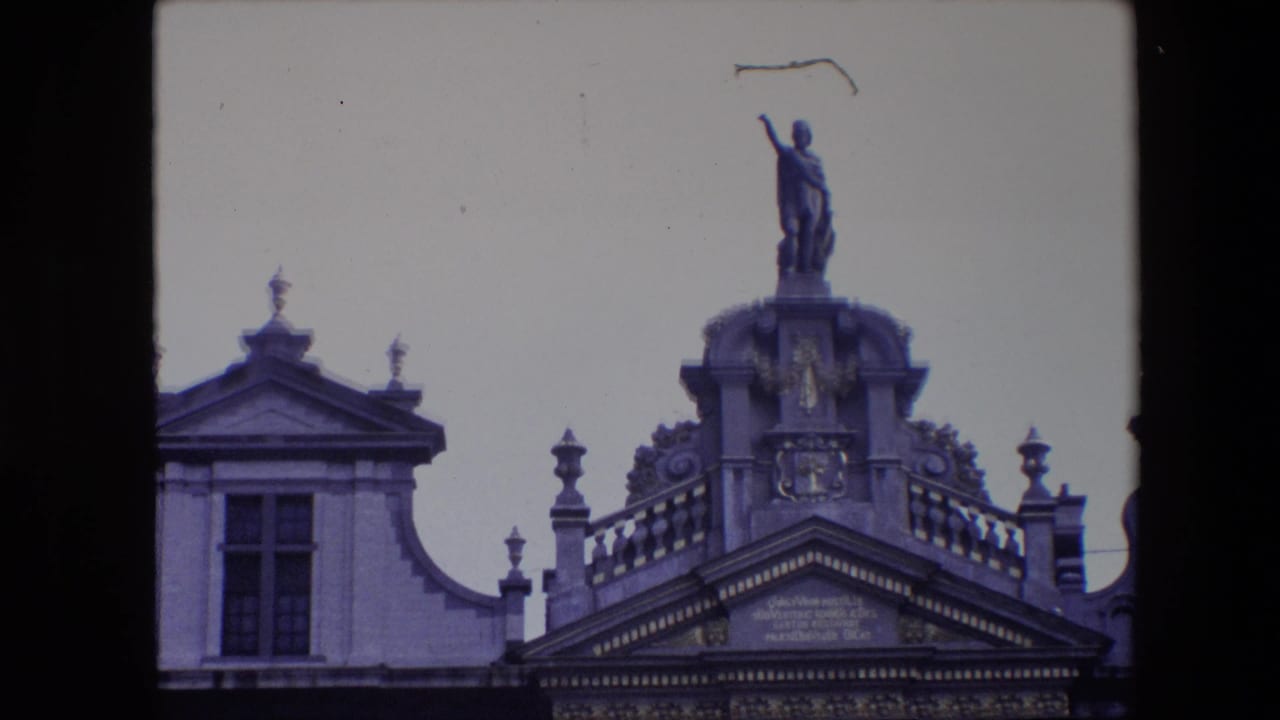 1967 Brussels 8mm frame showing the upper portion of an ornate historic facade with a rooftop statue, balustrade, pediment, and pale sky.