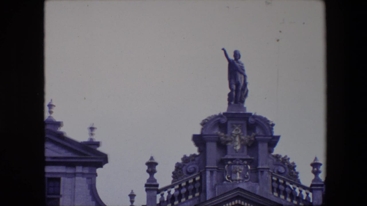 1967 Brussels 8mm frame with the camera closer on a robed rooftop statue standing above carved architectural ornament and a partial roofline.