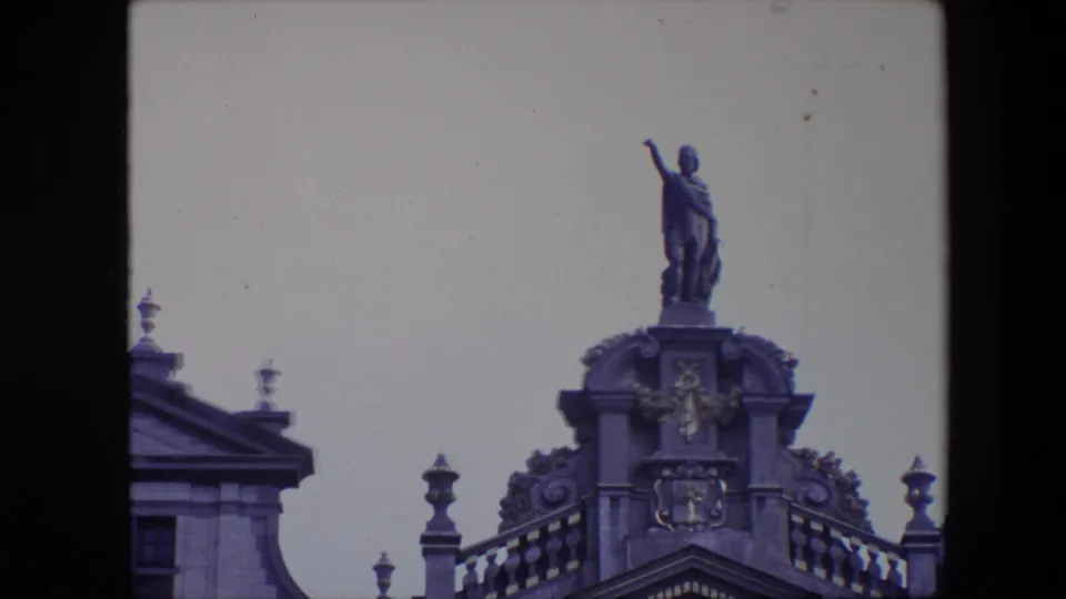 1967 Brussels 8mm frame with the camera closer on a robed rooftop statue standing above carved architectural ornament and a partial roofline.