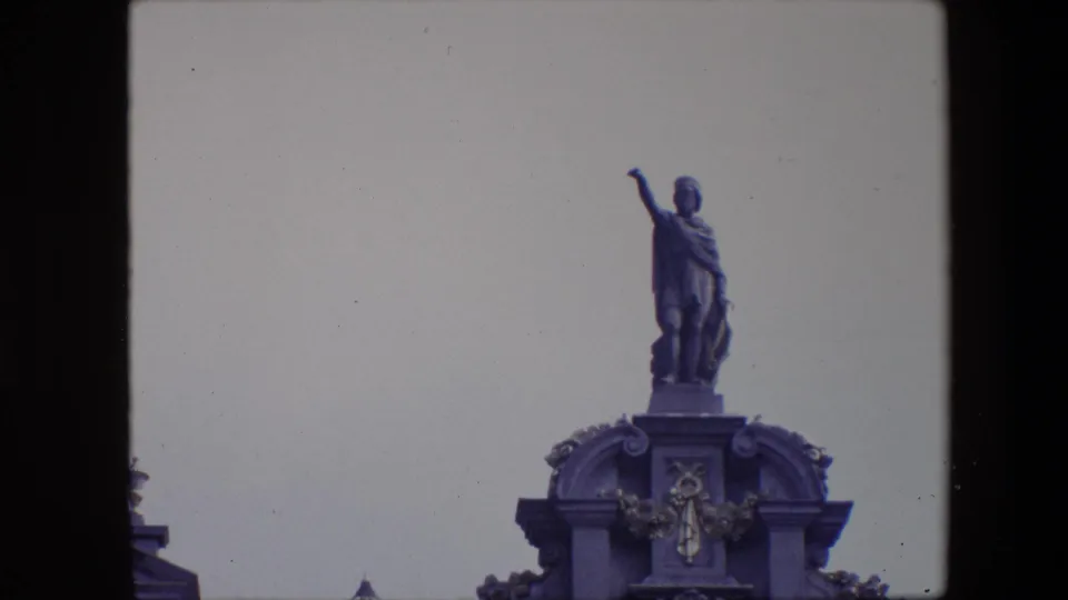 1967 Brussels 8mm frame centering the robed rooftop figure over an arched architectural crest, with the building cropped tightly against the sky.
