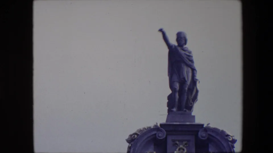 1967 Brussels 8mm frame showing a tight view of the robed rooftop statue with one raised arm, standing above cropped ornamental stonework against a pale sky.