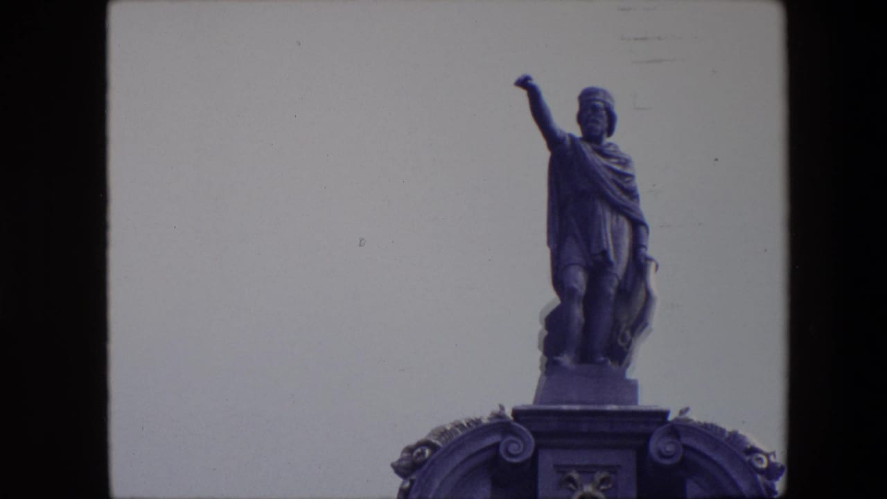 1967 Brussels 8mm frame showing a robed male statue with one arm extended, seen above a decorated pedestal against a pale sky.
