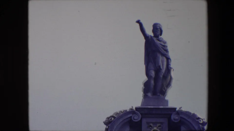 1967 Brussels 8mm frame showing a robed male statue with one arm extended, seen above a decorated pedestal against a pale sky.