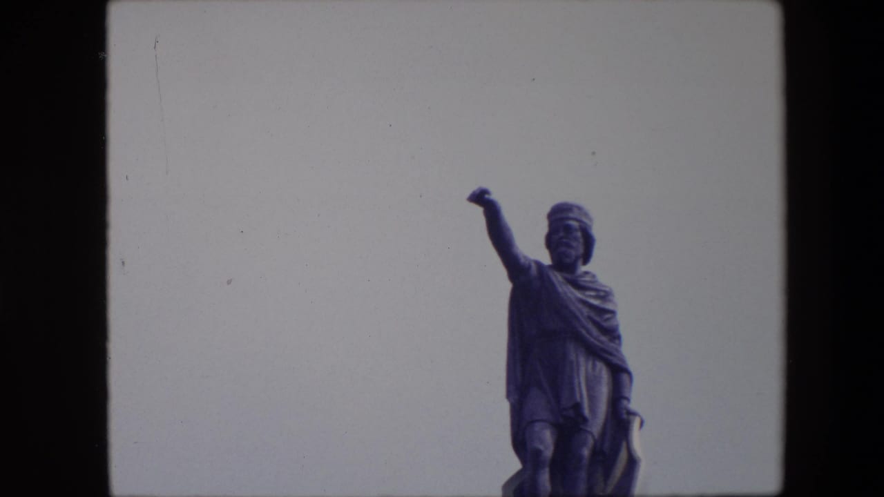 1967 Brussels 8mm frame with the robed statue shifted to the right side of the frame, its raised arm silhouetted against a pale sky.