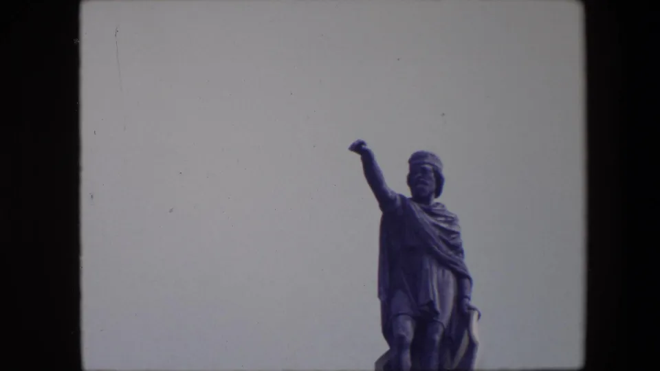 1967 Brussels 8mm frame with the robed statue shifted to the right side of the frame, its raised arm silhouetted against a pale sky.
