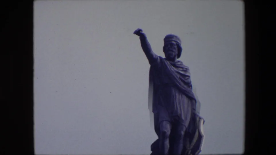 1967 Brussels 8mm frame presenting the robed male statue in closer right-side framing, with the extended arm, draped cloak, and lower figure visible against the sky.