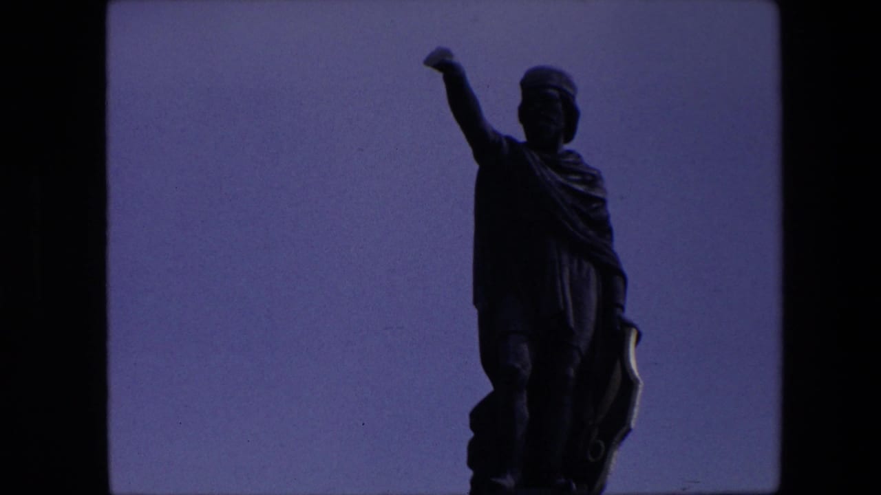 1967 Brussels home-movie frame showing a classical-looking statue in silhouette with one arm raised, bordered by dark 8mm film edges.
