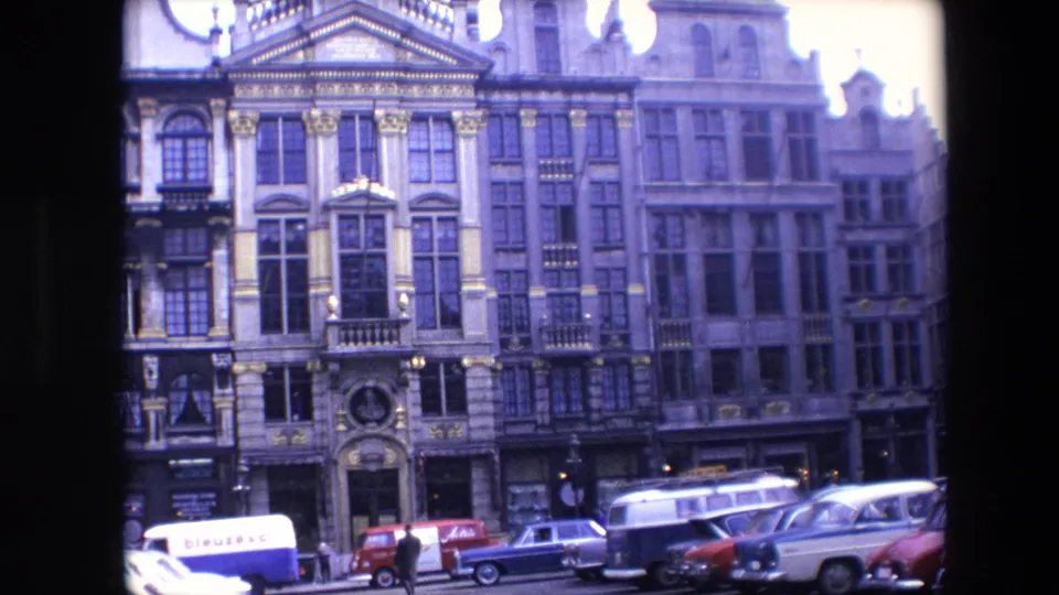 Street-level 1967 Brussels view of ornate historic facades, shopfronts, pedestrians, parked cars, and a white van with visible lettering.