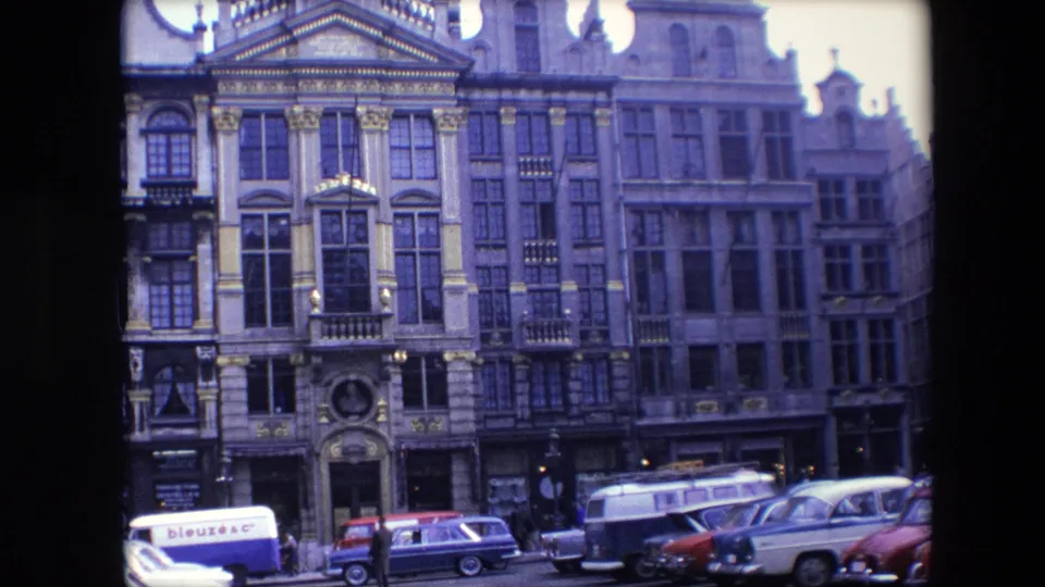 1967 Brussels home-movie frame showing decorated stone facades, large windows, street-level shops, pedestrians, and mid-century vehicles below.