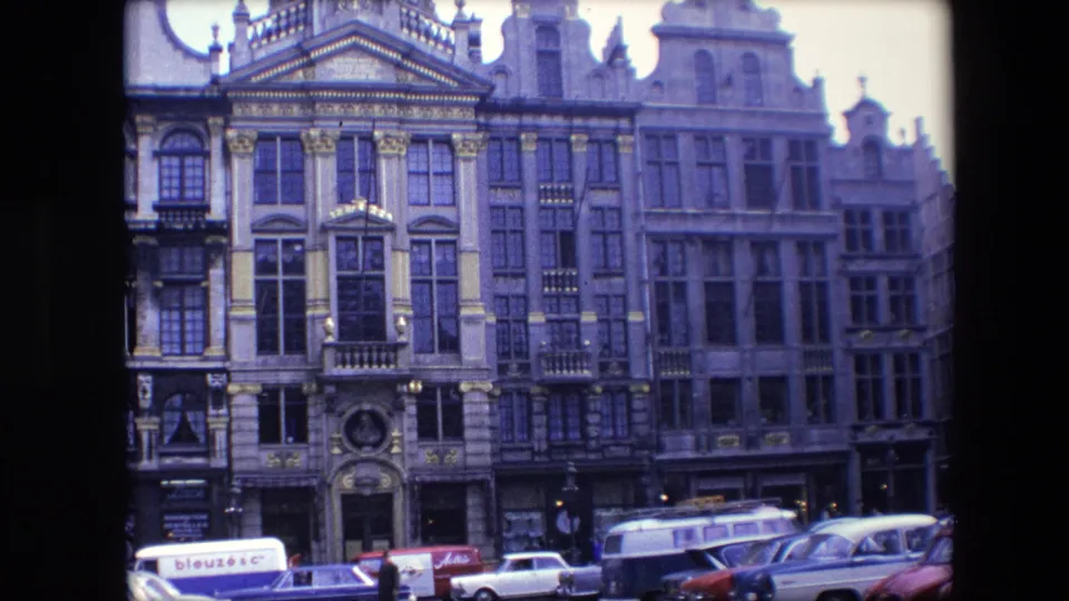 Handheld 1967 Brussels street view with ornate multi-story buildings, dark film borders, pedestrians, and several parked 1960s cars.