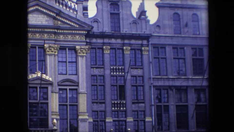 1967 Brussels home-movie frame showing an ornate historic facade with gilded details, tall multi-paned windows, and pale classical columns.