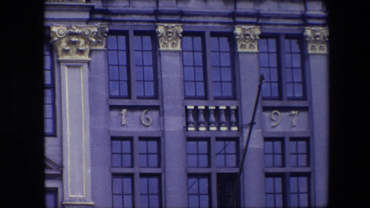 1967 Brussels 8mm frame of an upper historic facade with ornate capitals, dark grid windows, balcony-like stone detail, date numerals, and an empty flagpole.