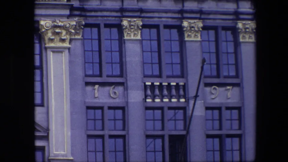 1967 Brussels 8mm frame of an upper historic facade with ornate capitals, dark grid windows, balcony-like stone detail, date numerals, and an empty flagpole.