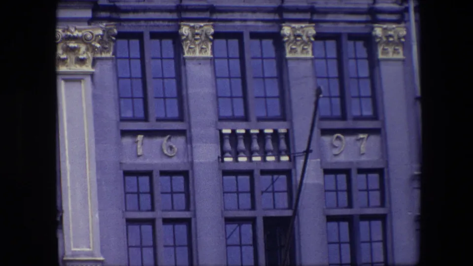 1967 Brussels 8mm frame of a historic building facade with repeated dark window grids, carved capitals, pale pilaster panels, date numerals, and an angled empty flagpole.