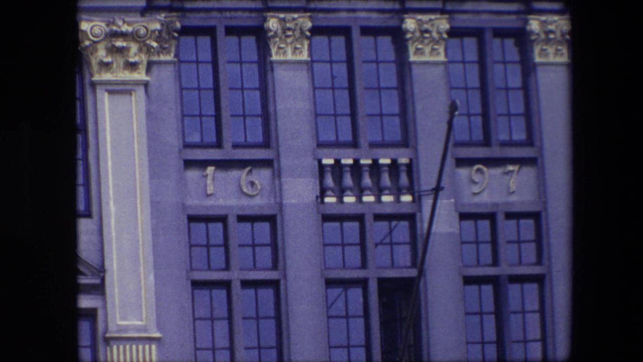 1967 Brussels home-movie frame showing an ornate historic facade with tall dark windows, gilded architectural details, and the number 1697 above lower windows.