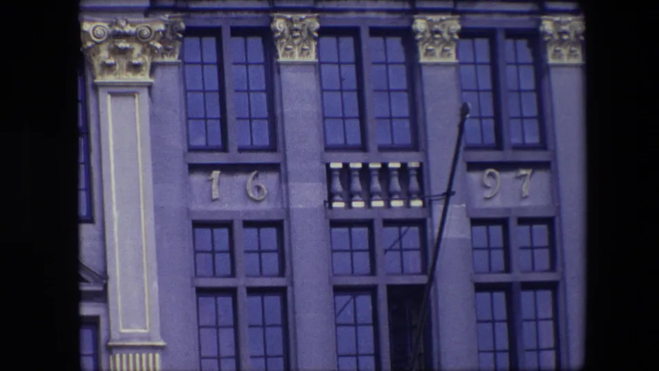 1967 Brussels home-movie frame showing an ornate historic facade with tall dark windows, gilded architectural details, and the number 1697 above lower windows.