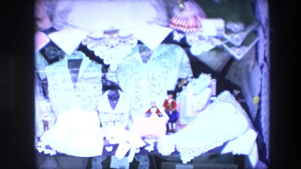 1967 Brussels home-movie frame showing a bright lace and textile display with white garments, small dolls, and layered decorative fabric objects.