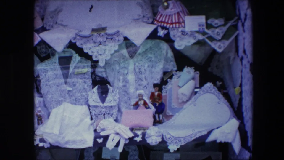 1967 Brussels 8mm frame showing white lace garments and accessories arranged with small folk-costume dolls in a close display.