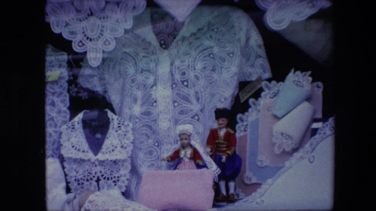 1967 Brussels home-movie frame with intricate white lace garments, small traditional-costume dolls, and folded pastel accessories on display.