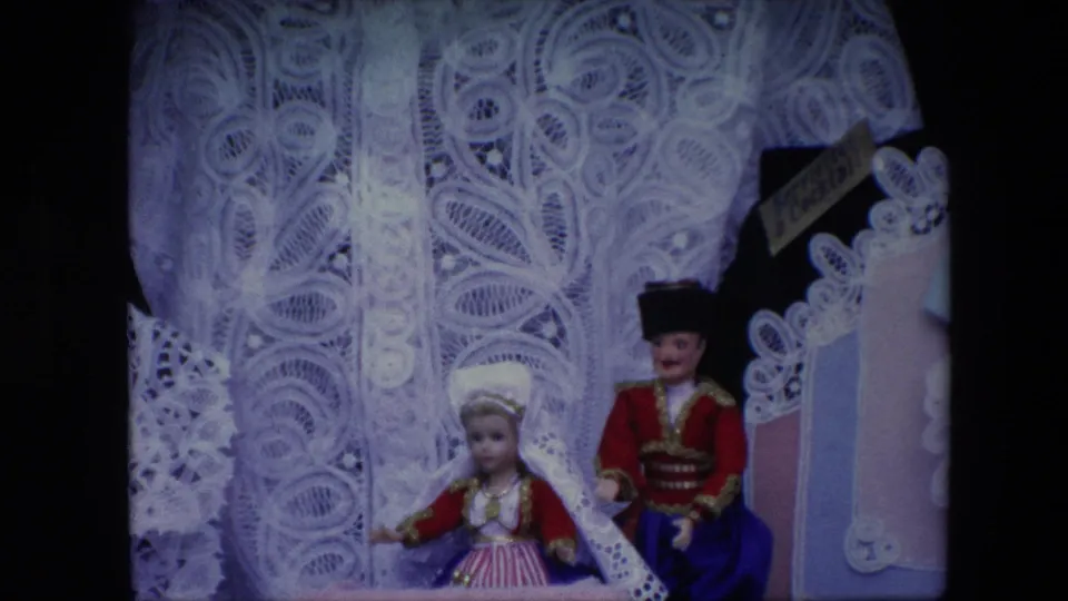 1967 Brussels 8mm close-up emphasizing the patterned white lace garment and two small folk-costume dolls at the base of the display.