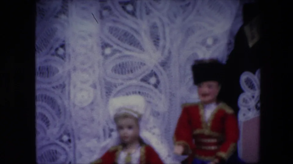 Soft 1967 Brussels 8mm close-up of white lace patterning with two small traditional-costume dolls blurred in front.