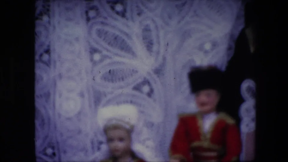 1967 Brussels 8mm frame showing two fancy dressed dolls, one with a white headpiece and one in a red uniform-style costume, set against a pale decorative filigreed backdrop.