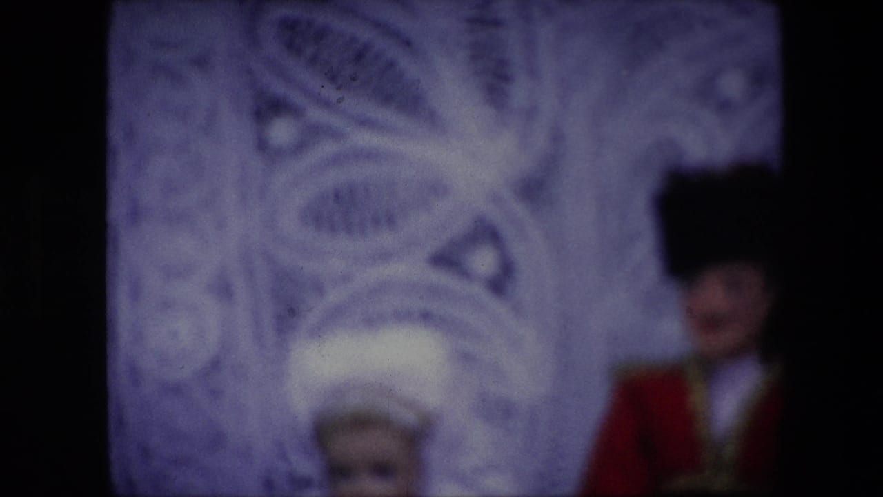 Soft 1967 Brussels home-movie frame of two fancy dressed dolls, with the male doll in red and black at right and the white headpiece of the other doll partly visible near the lower center.
