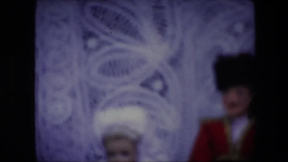 Soft 1967 Brussels home-movie frame of two fancy dressed dolls, with the male doll in red and black at right and the white headpiece of the other doll partly visible near the lower center.
