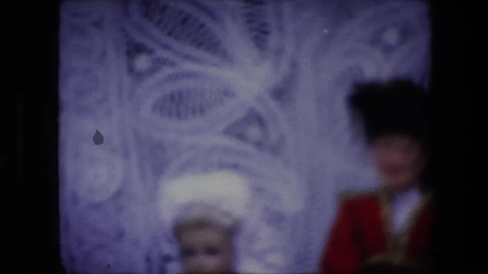 1967 Brussels 8mm frame with a blurred view of the white headpiece and red uniform-style doll costume against a pale patterned filigree backdrop.