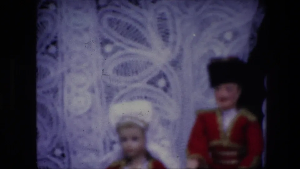 1967 Brussels 8mm frame showing the two costumed display dolls, including a red uniform-style outfit and white headpiece, beside a dark vertical border and ornate pale backdrop.