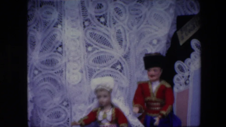1967 Brussels home-movie frame showing two porcelain dolls in traditional-style red and gold clothing against a white lace display backdrop.