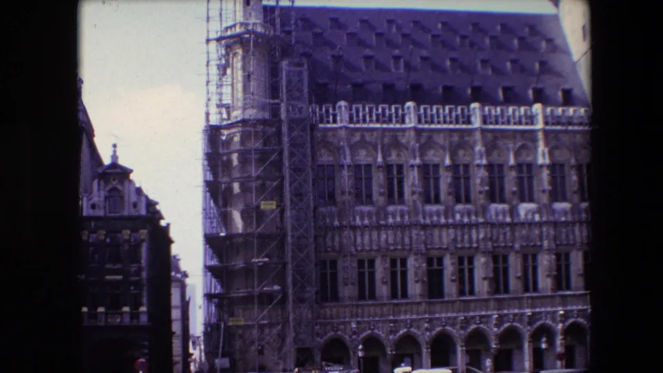 1967 8mm view in Brussels showing an ornate historic stone building with scaffolding, arched openings, tall windows, and a steep dark roof.