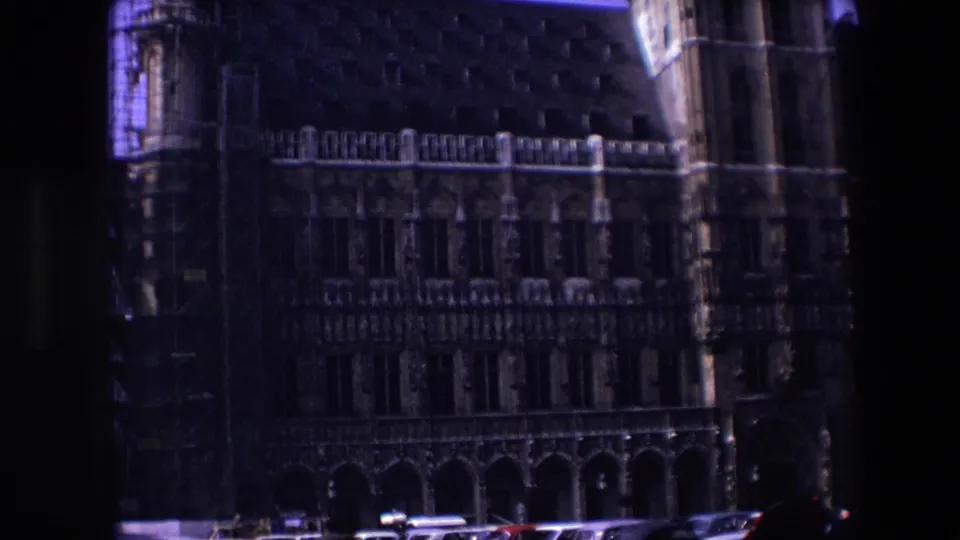 Underexposed 1967 Brussels home-movie frame showing the Gothic facade, left-side scaffolding, roofline details, and vehicles along the street.