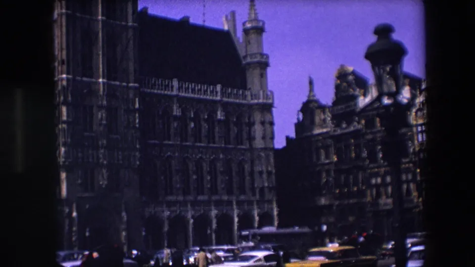 1967 Brussels frame from a wider angle, showing the historic Gothic building receding along the square, cars at street level, and a dark street lamp in the foreground.