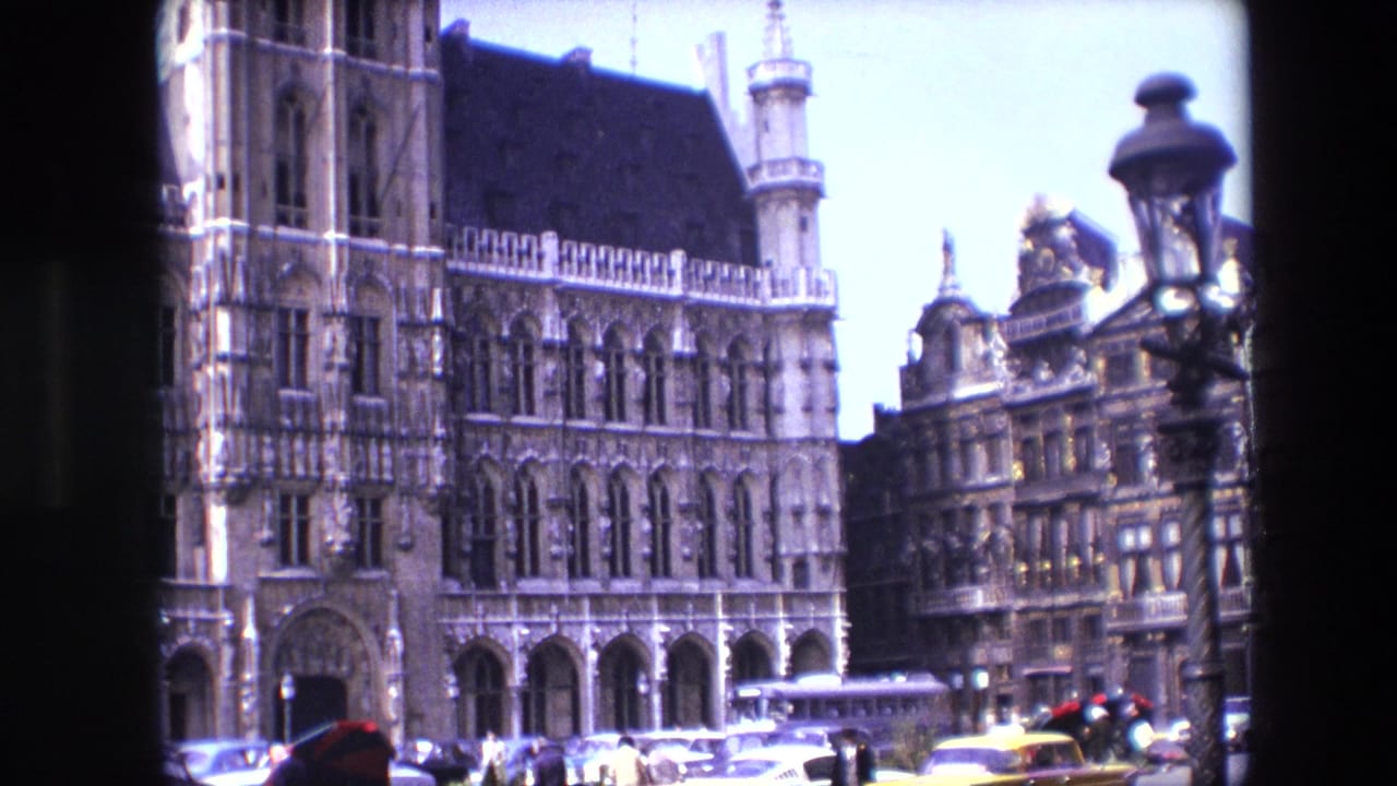 1967 Brussels home-movie frame showing the ornate Town Hall facade above a busy square with cars, yellow taxis, pedestrians, a bus, and a street lamp.